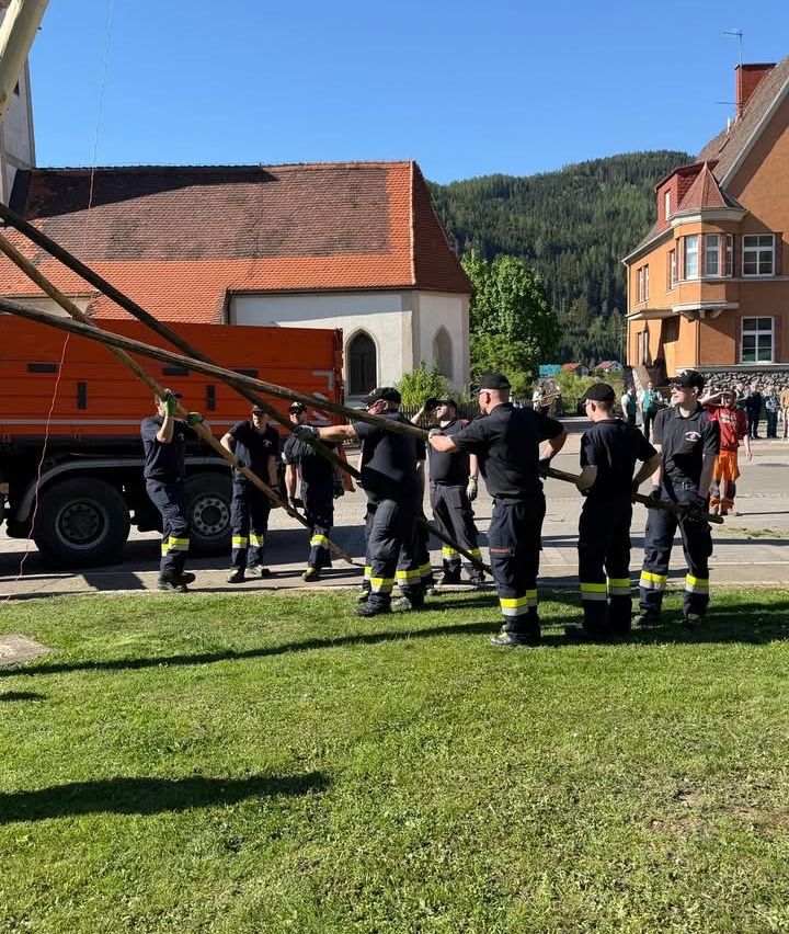 Eine Gruppe von Feuerwehrleuten hält große Holzstangen in einem Park. Ein Lkw ist hinter ihnen geparkt. Im Hintergrund befinden sich Gebäude und Bäume.