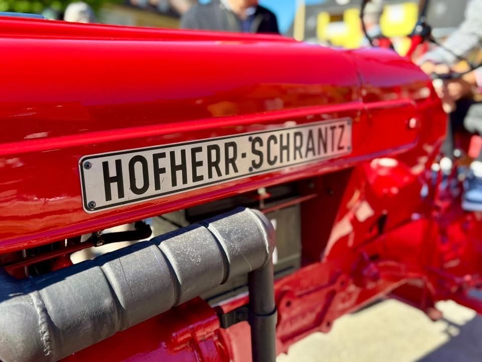 Ein roter Vintage-Traktor mit einem Namensschild, das 'HOFHERR-SCHRANTZ' liest, an der Vorderseite befestigt. Der Traktor hat einen metallischen Rohr, der an seiner Seite angeschlossen ist.