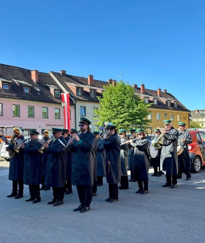 Ein Blechbläserensemble in dunklen Mänteln steht an einem sonnigen Tag in einer Reihe, mit Gebäuden und einem roten Auto im Hintergrund.