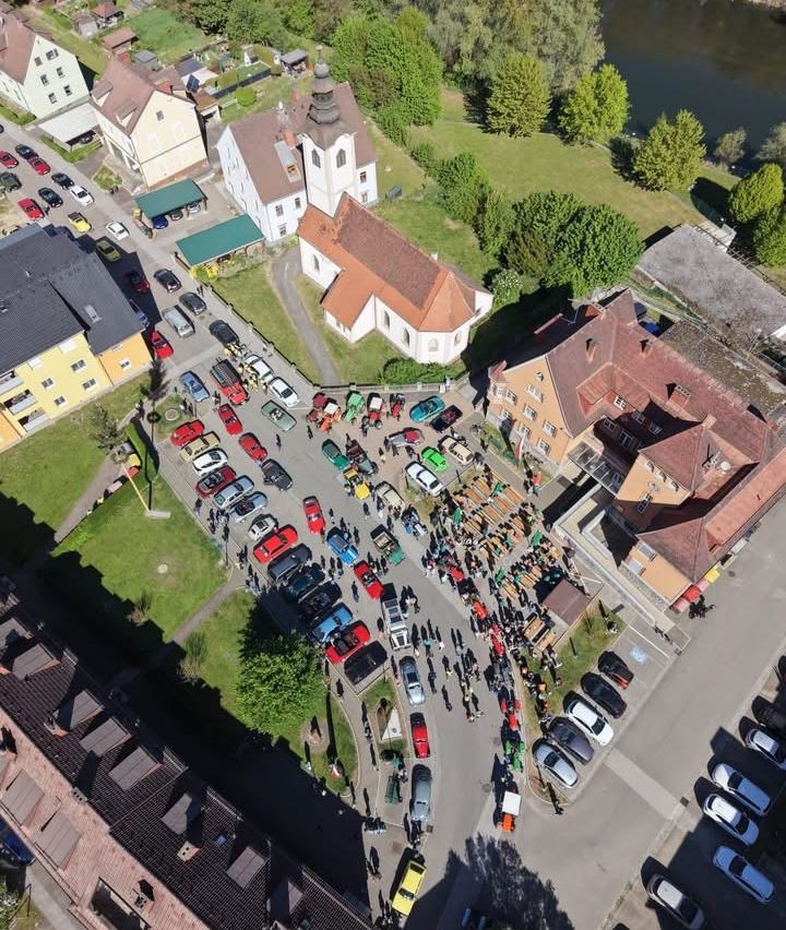 Ein Luftbild einer Stadt mit vielen auf der Straße geparkten Autos. Es gibt Menschen, die auf dem Bürgersteig gehen. Es gibt Gebäude, Bäume und einen Rasen.