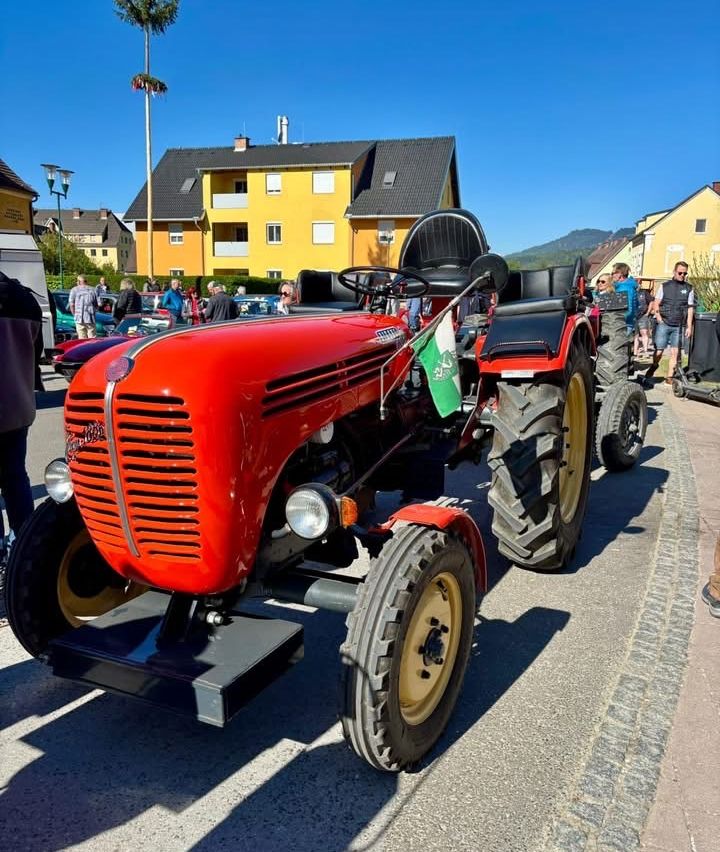 Ein roter Oldtimer-Traktor steht am Straßenrand geparkt, mit Menschen und Autos im Hintergrund. Der Traktor hat eine grüne Flagge, die daran befestigt ist.