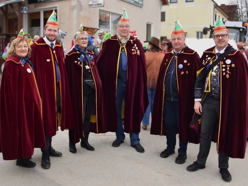 Eine Gruppe von Menschen in festlichen roten Umhängen und Kegelhüten, die in einer Reihe vor einem Gebäude stehen.