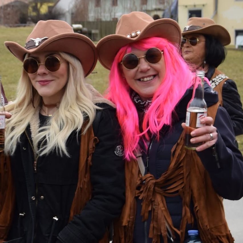 Zwei Frauen in Cowboy-Huten mit rosa Haaren und Sonnenbrillen lächeln und halten Flaschen. Dahinter eine Frau in einem braunen Hut und einer Brille. Ein Gebäude und Gras sind im Hintergrund.