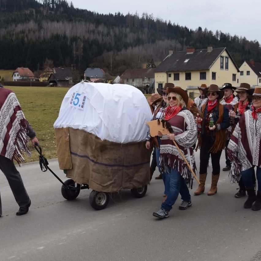Eine Gruppe von Menschen in Cowboy-Kleidung marschiert bei einer Parade und hält einen Holzwagen mit der Nummer 5. Dahinter hält ein Mann in einem roten und weißen Poncho einen Stock. Die Parade findet auf einer Straße statt, mit Häusern im Hintergrund.