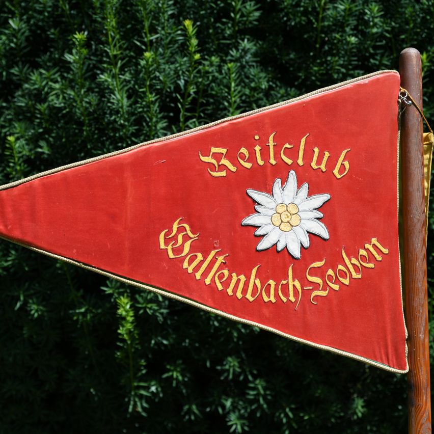 A red triangular flag with golden text reading 'Reitclub Falkenbach-Sebern' and a white flower emblem on it. The flag is mounted on a wooden pole.