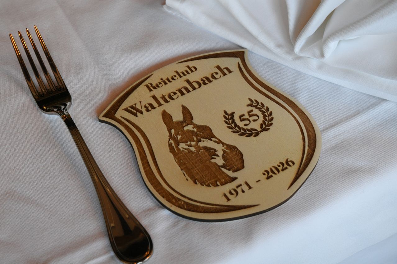 A wooden plaque with a horse logo and the text 'Reitclub Waltenbach', dated from 1971 to 2026, placed on a white cloth next to a silver fork.