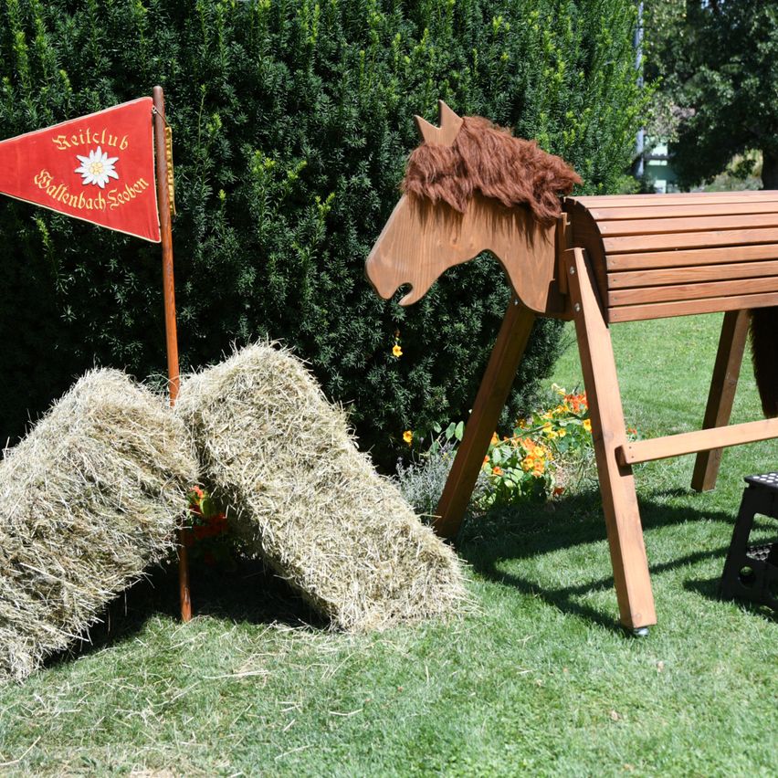 A wooden horse with a red flag labeled 'Reitclub Wallenbach Sachsen' stands on a grassy field with hay bales and a wooden fence in the background.