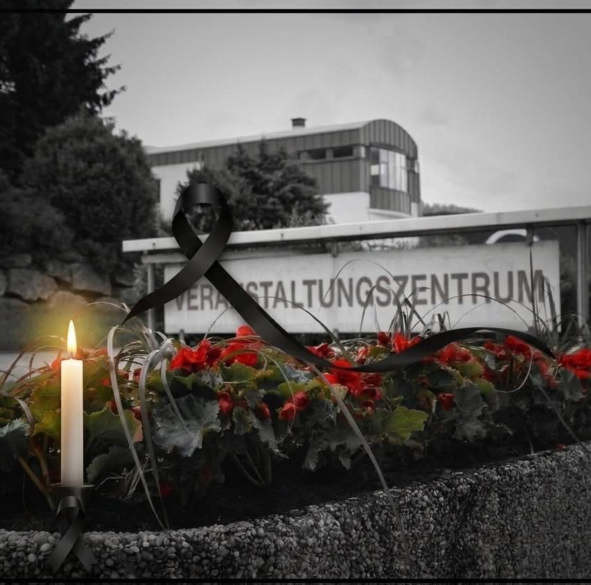 Ein schwarz-weißes Bild eines Denkmals mit einer Kerze, Blumen und einem schwarzen Band, das an ein Gebäude mit der Aufschrift 'Veranstaltungszentrum' gebunden ist.