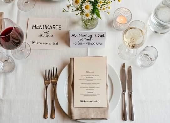 Ein Tisch mit weißer Tischdecke, einer Menükarte, Weingläsern und Besteck. In der Mitte steht eine Vase mit Gänseblümchen. Die Menükarte lautet 'MENÜKARTE' und 'Vaz Niklasdorf'. Der Tisch ist für eine Mahlzeit vorbereitet.