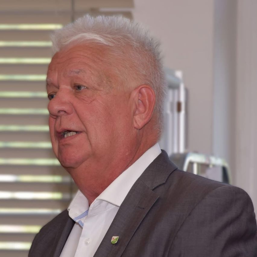 Ein älterer Mann mit weißen Haaren, der einen grauen Anzug und ein weißes Hemd trägt, steht und spricht.