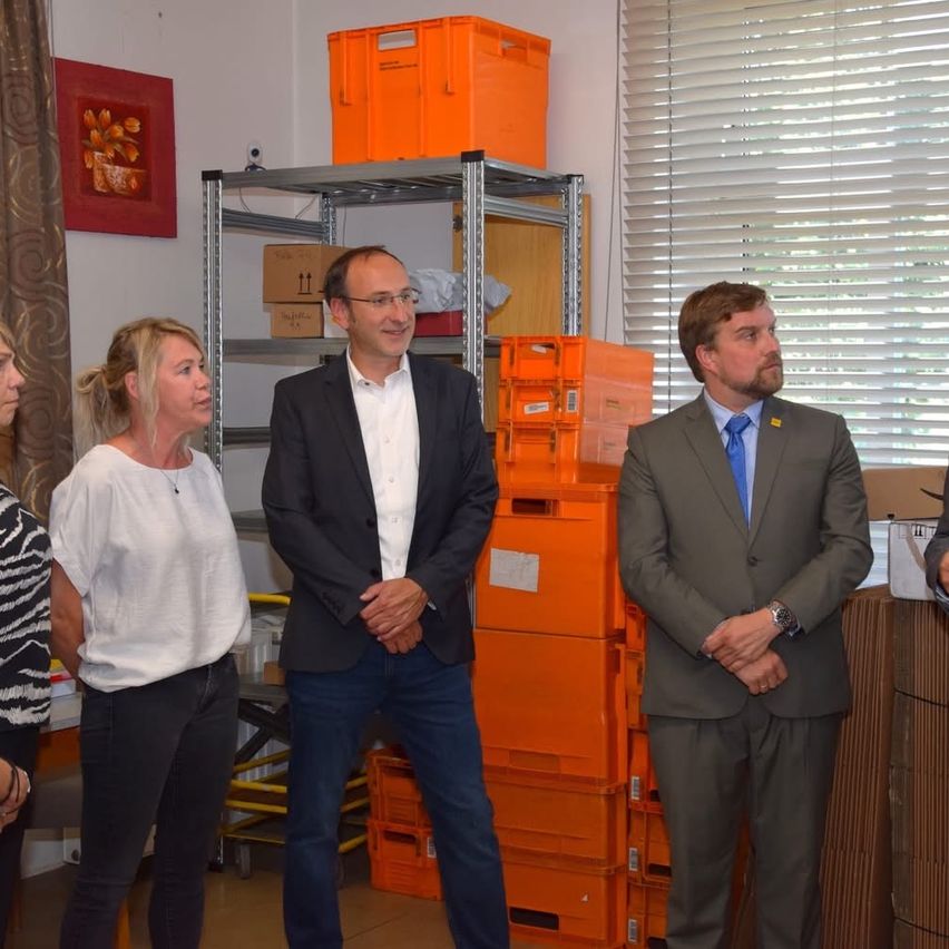 Drei Personen stehen in einem Raum mit orangefarbenen Kisten hinter ihnen. Ein Mann in einem Anzug steht zwischen zwei Frauen.