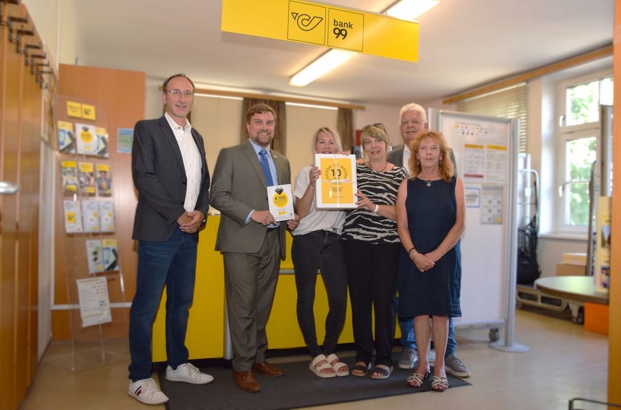 Eine Gruppe von Menschen steht vor einem gelben Hintergrund mit einem Bank 99-Schild und hält Urkunden. Sie sind formell gekleidet und scheinen eine Errungenschaft zu feiern.