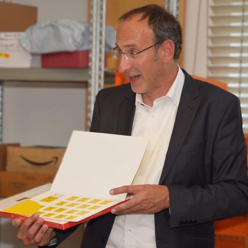 Ein Mann in einem Anzug hält eine Mappe mit gelben Aufklebern. Er steht in einem Raum mit Regalen und Kisten.