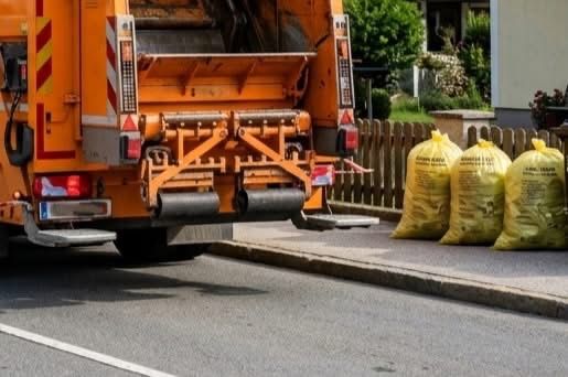 Ein Müllwagen steht am Straßenrand, mit drei gelben Müllsäcken in der Nähe. Der Wagen ist orange mit reflektierenden Streifen. Dahinter befindet sich ein Holzzaun und ein Gebäude.