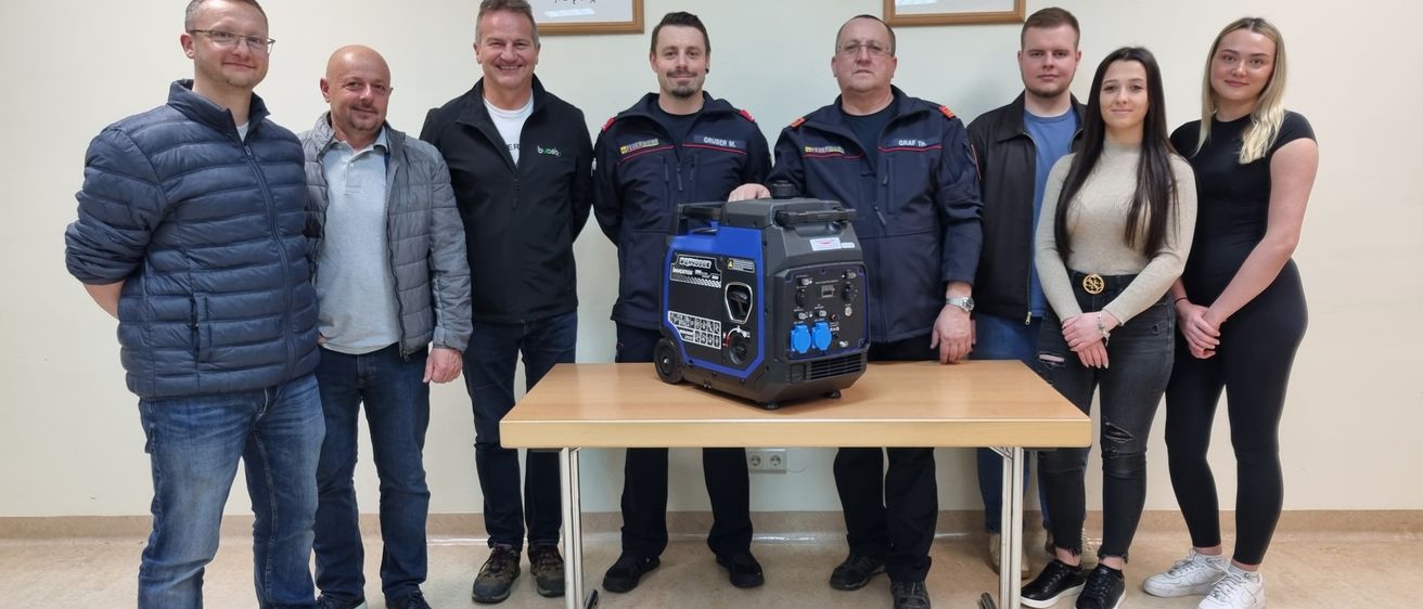 Acht Personen stehen um einen Tisch mit einem Generator in der Mitte. An der Wand dahinter hängen drei gerahmte Bilder.