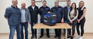 Acht Personen stehen um einen Tisch mit einem Generator in der Mitte. An der Wand dahinter hängen drei gerahmte Bilder.