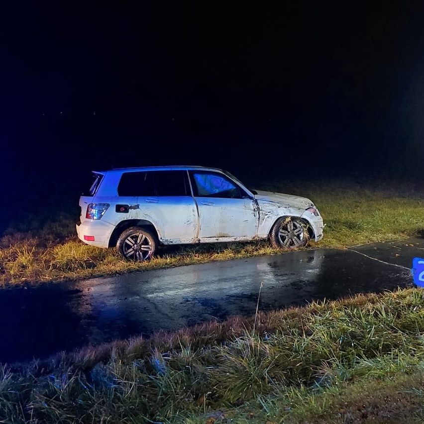 Ein weißer SUV ist auf der nassen Straße am Straßenrand geparkt, mit Schäden an der Rückseite, bei Nacht.