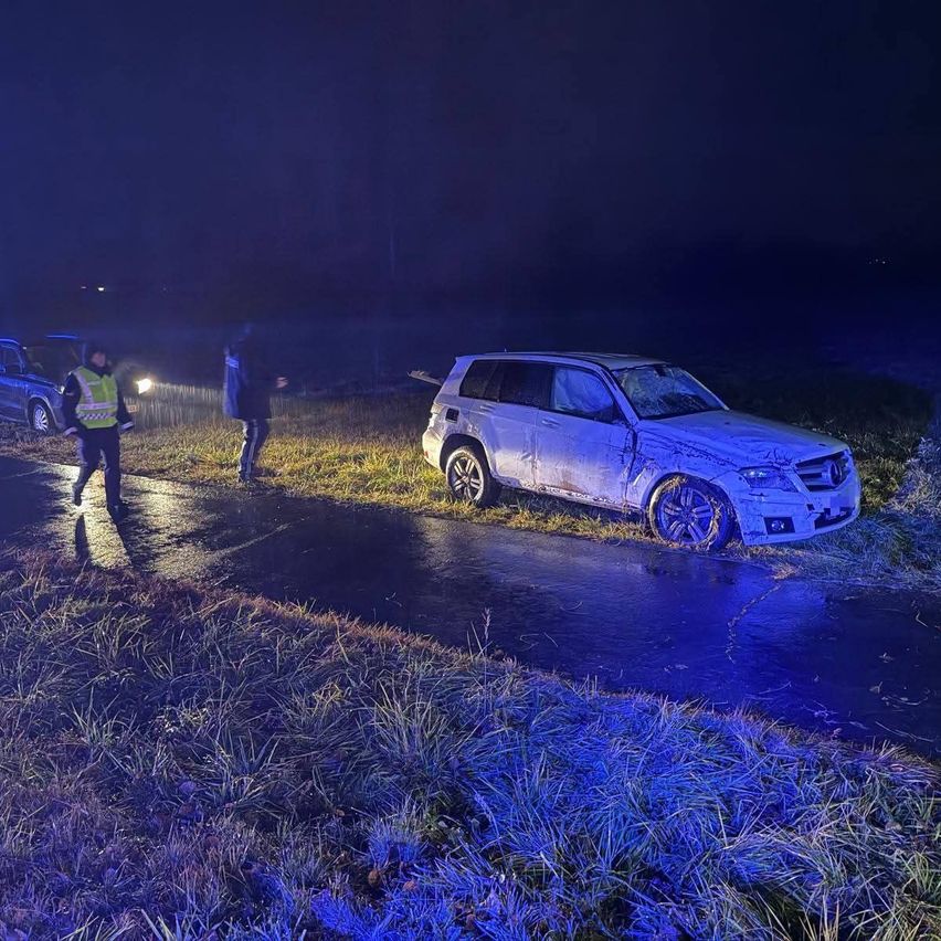Zwei Personen, eine in einer reflektierenden Weste, stehen neben einem beschädigten weißen SUV auf einer nassen Straße bei Nacht. Die Front des SUV ist stark beschädigt.