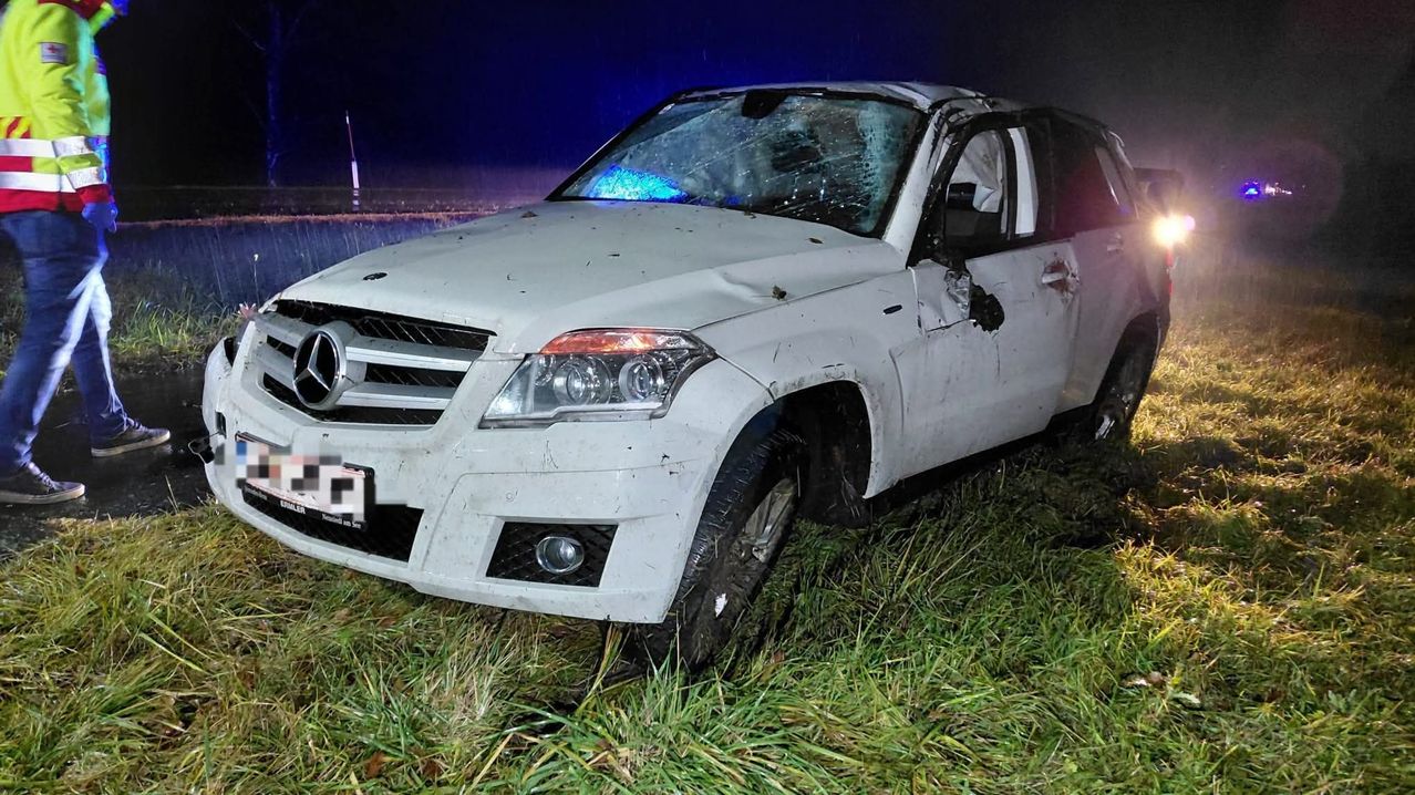 Ein weißer Mercedes-Benz SUV ist auf einem Grasfeld bei Nacht zerstört, mit stark beschädigter Front, eingeschalteten Scheinwerfern und blauen Lichtern, die die Szene beleuchten.
