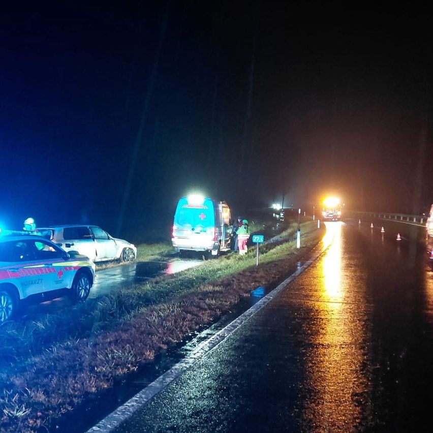 Eine nächtliche Szene mit einem Rettungswagen am Straßenrand. Ein Auto ist auf der linken Seite geparkt, und ein weiteres Fahrzeug ist auf der Straße. Verkehrskegel sind am Straßenrand aufgestellt.