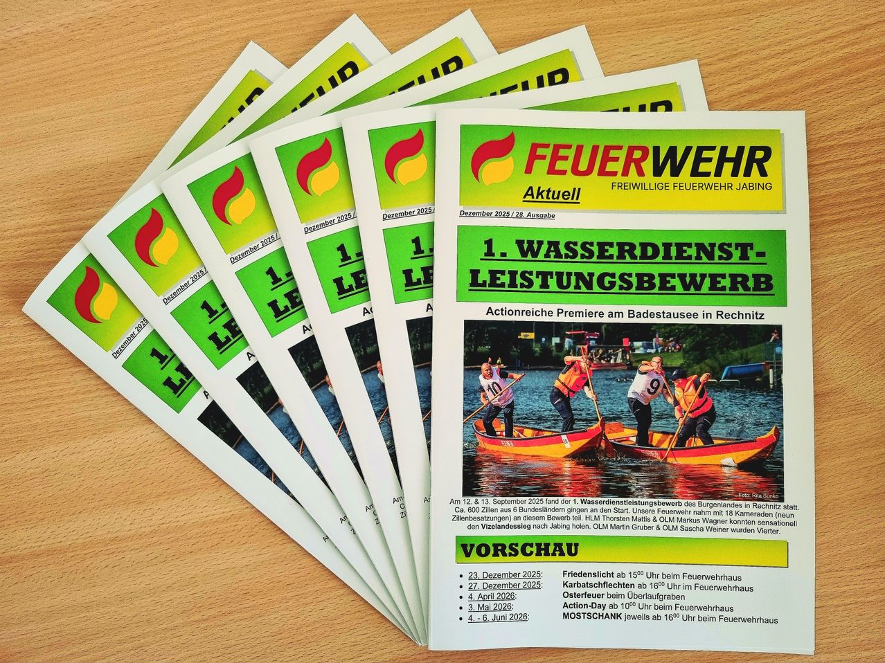 Stapel von gedruckten Flyern auf einer Holzoberfläche. Der oberste Flyer trägt den Titel 'FEUERWEHR Aktuell' und zeigt ein Bild von Menschen in Booten. Es enthält Auflistungen von Veranstaltungen und Terminen, darunter '1. Wasserdienstleistungsbewerb' und 'Aktionreiche Premiere'.
