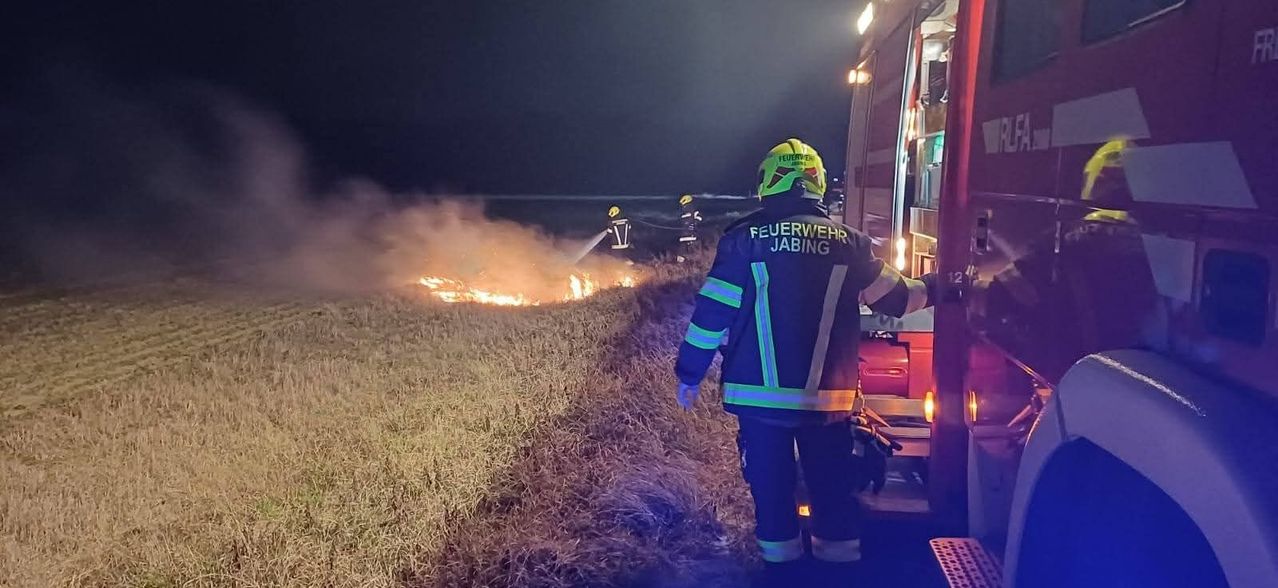 Firefighters in protective gear are battling a fire on a grassy field under a dark night sky.