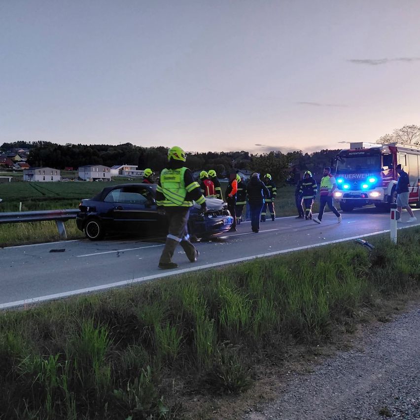 Emergency responders in yellow vests attend to a wrecked car on the road, with a fire truck nearby.