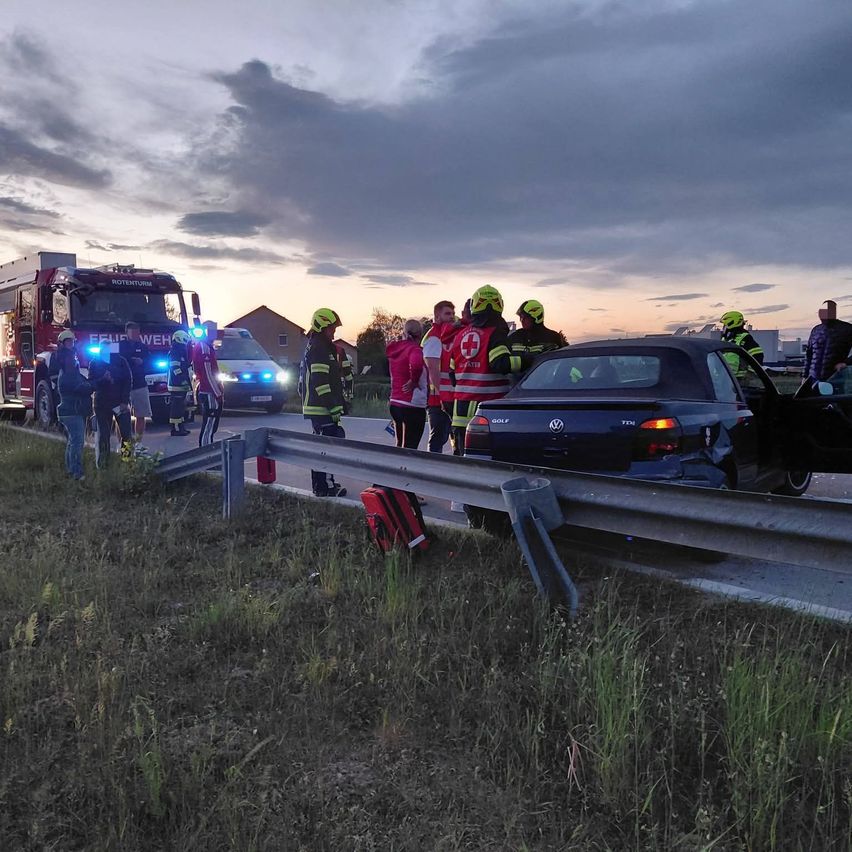 An emergency vehicle with flashing lights is parked on the side of the road. Several people are gathered around the car, with some wearing helmets and vests.