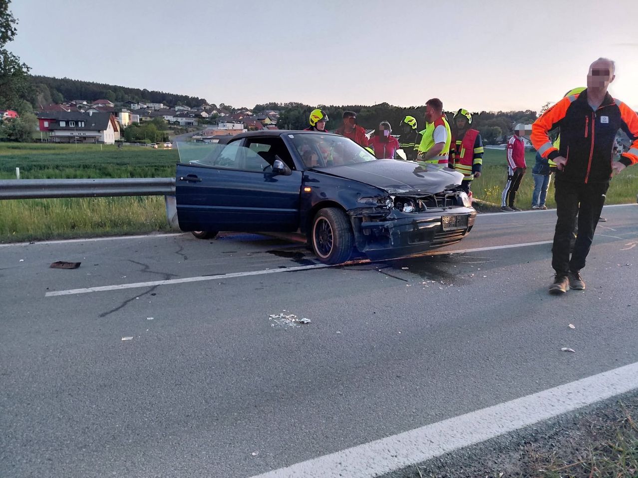 A blue car with a damaged front is parked on the road. Several people in safety gear are around it. A person stands at the roadside, wearing an orange vest.