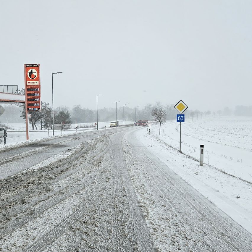Verschneite Straße mit einer Tankstelle links und einem Geschwindigkeitsschild rechts. Einige Fahrzeuge fahren auf der Straße.