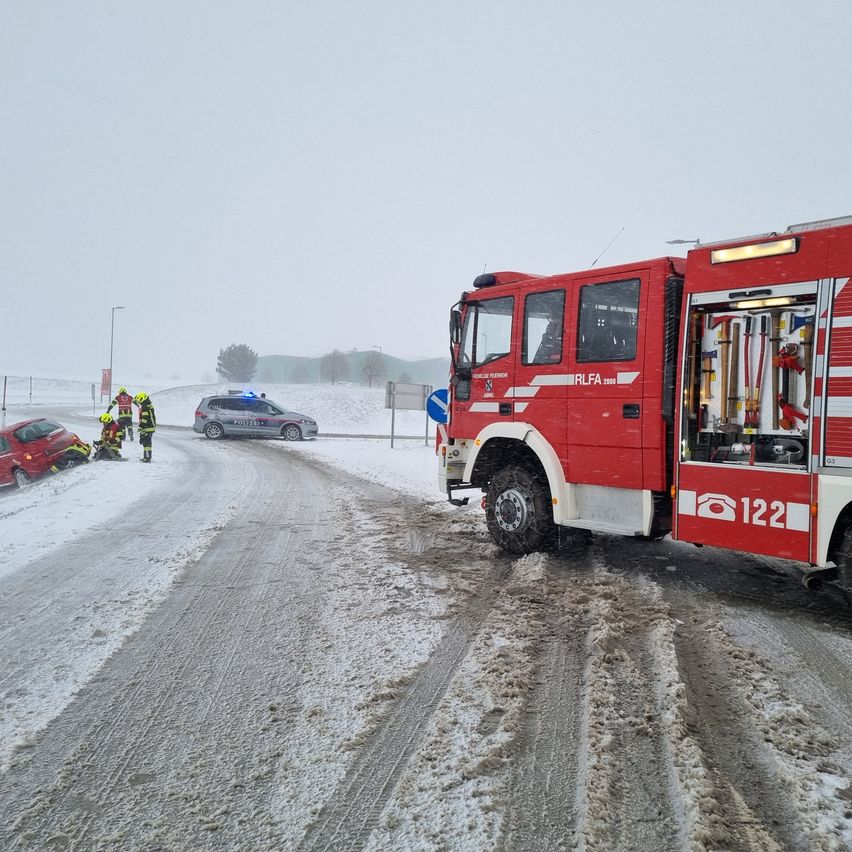 Ein roter Feuerwehrwagen ist auf einer verschneiten Straße geparkt, mit zwei Personen und einem Auto an der Seite. Ein Polizeiauto ist ebenfalls in der Nähe geparkt.