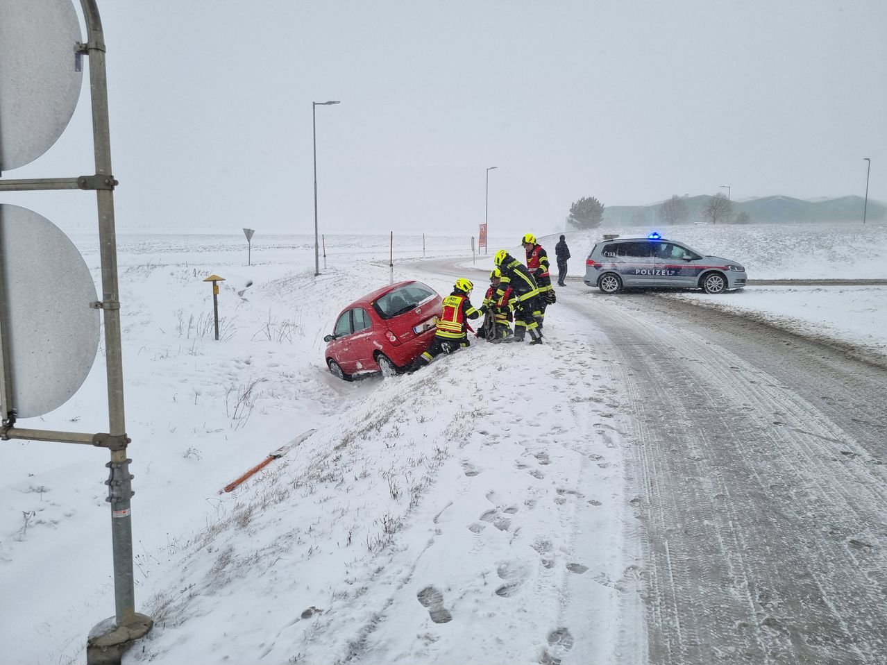 Feuerwehrleute in gelber und roter Ausrüstung kümmern sich um ein rotes Auto in einem verschneiten Gebiet. Ein Polizeifahrzeug ist in der Nähe geparkt.