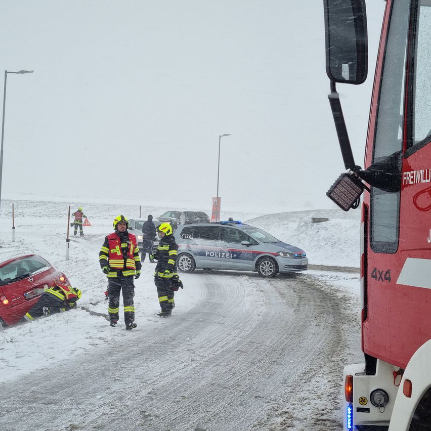 Zwei Feuerwehrleute stehen auf einer verschneiten Straße, in der Nähe eines demoliert roten Autos und eines Polizeiwagens. Sie tragen reflektierende Kleidung und Helme. Schnee bedeckt den Boden, und ein roter Feuerwehrwagen ist in der Nähe.