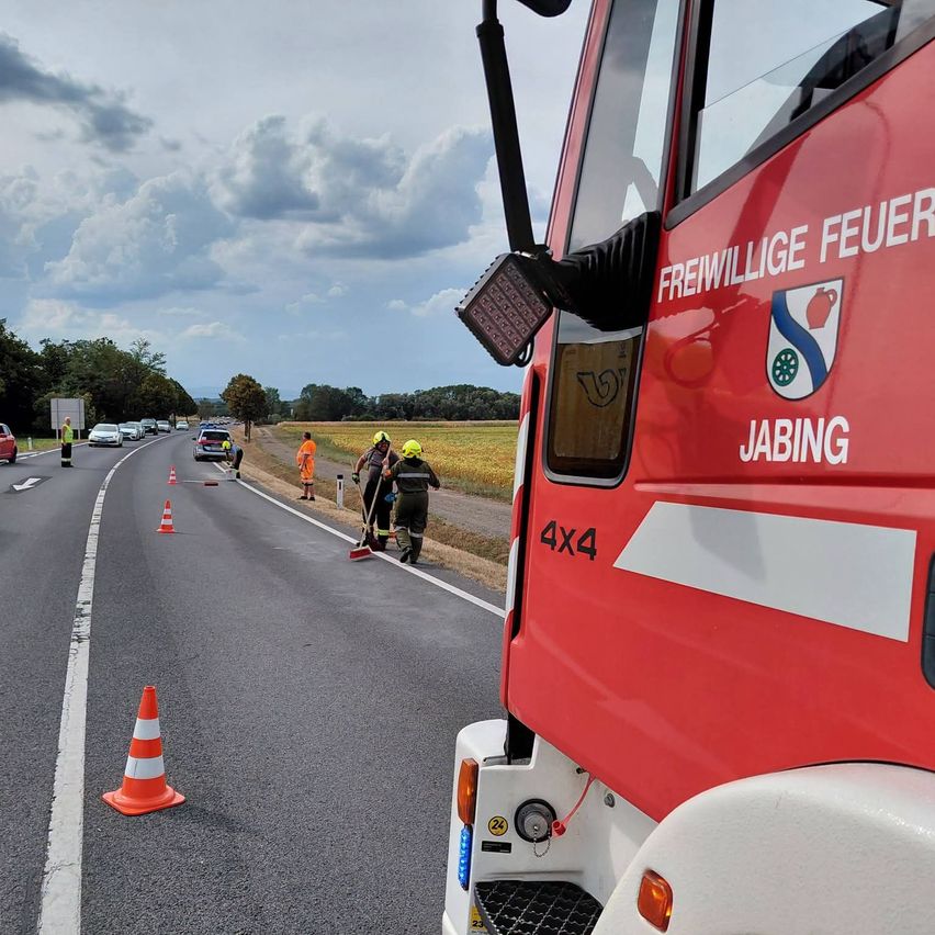 Ein roter Feuerwehrwagen steht am Straßenrand mit Verkehrskegel und Personen in Schutzkleidung. Der Wagen trägt die Aufschrift 'Freiwillige Feuerwehr Jabing' und '4x4'.