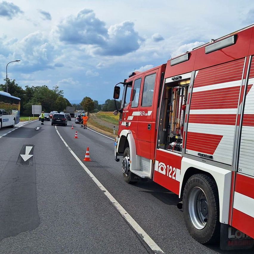Ein roter Feuerwehrwagen steht mit offener Tür am Straßenrand. Verkehrskegel sind auf der Straße platziert, und ein Bus ist auf der linken Seite geparkt. Eine Person steht in der Nähe der Verkehrskegel.