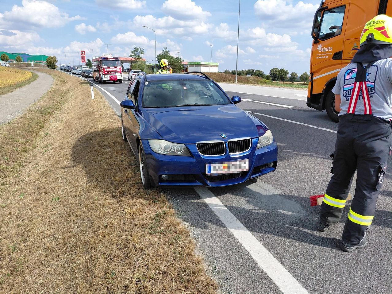 Ein blauer BMW ist am Straßenrand geparkt, in der Nähe befinden sich Rettungsfahrzeuge. Ein Feuerwehrmann steht neben dem Auto.