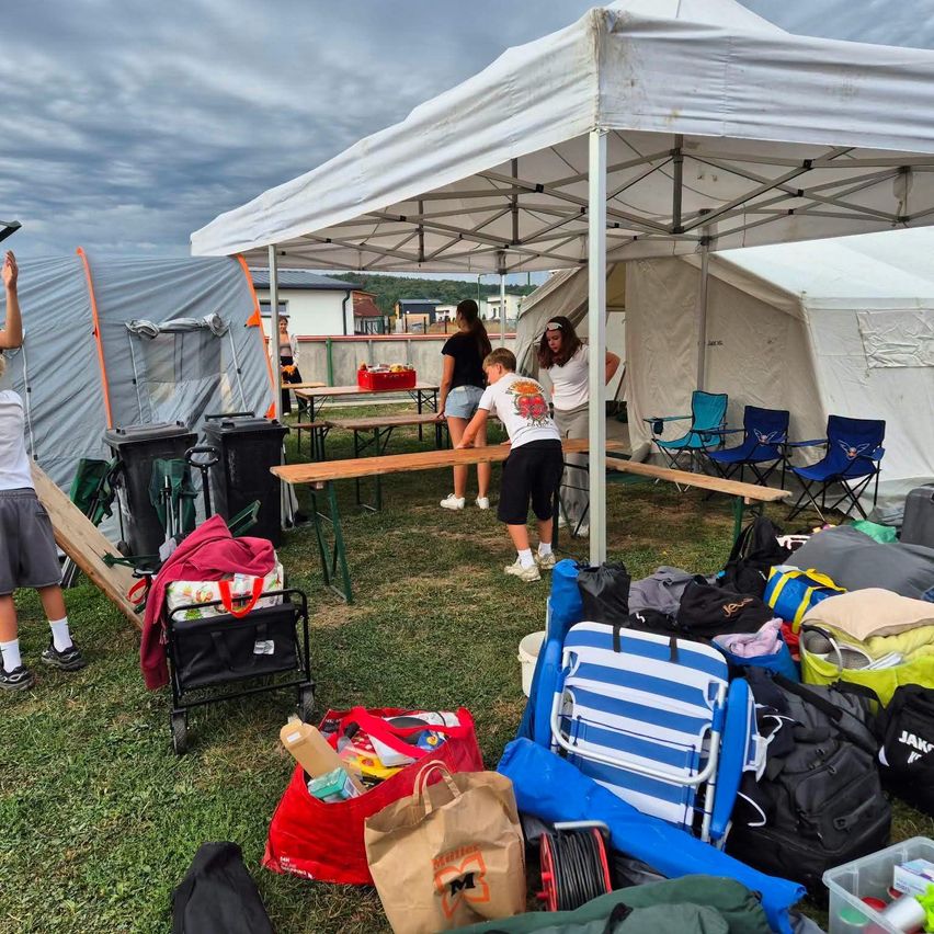 A group of people are setting up a tent and arranging tables and chairs on a grassy field. There are various items and bags scattered around.