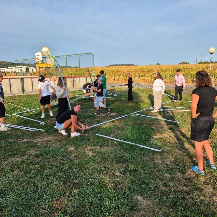 A group of people are assembling a metal structure on a grassy field with a cornfield in the background.