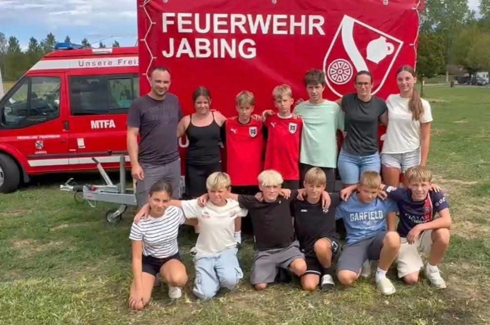 A group of people, including children and adults, are posing for a photo in front of a red banner with the words 'Feuerwehr Jabing' and a fire truck in the background.