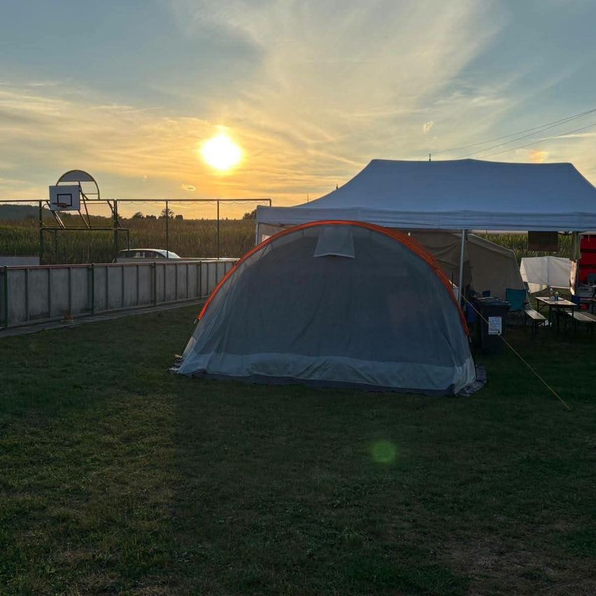 A tent set up in a grassy field with a large white tent behind it, a basketball hoop on a fence, and a sunset in the background.