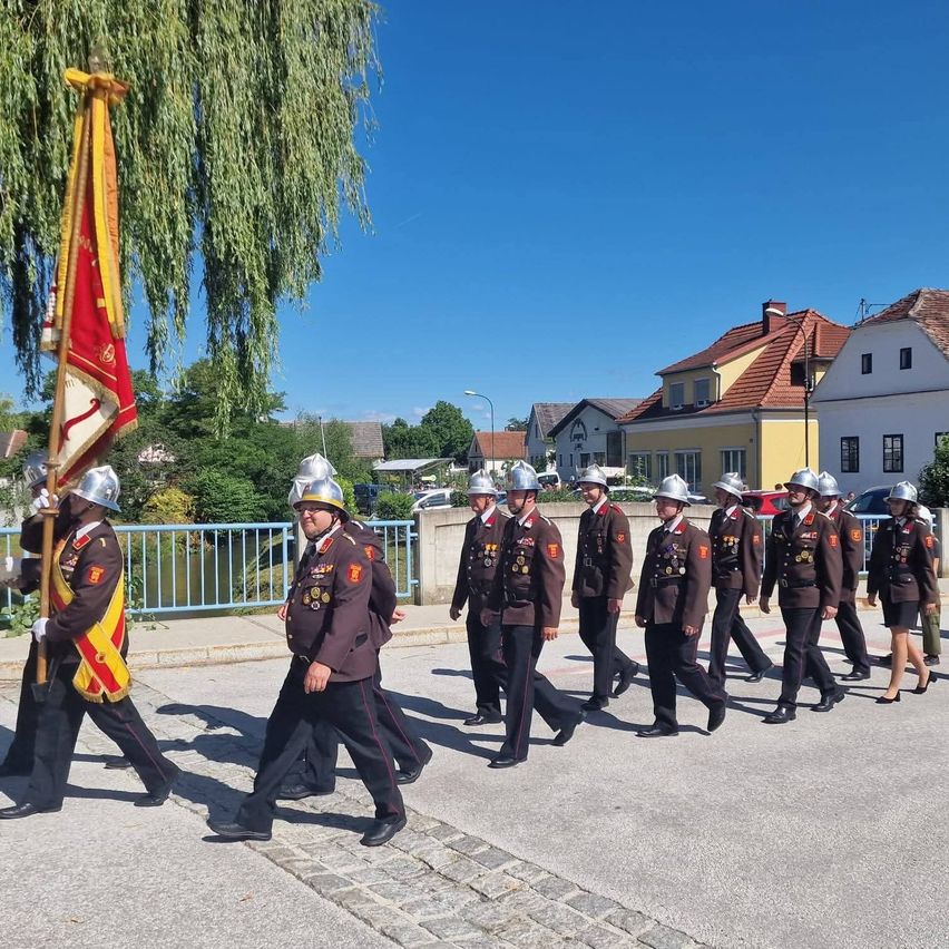 A marching band in military uniforms is parading down a street, with one member carrying a flag. Buildings line the street.