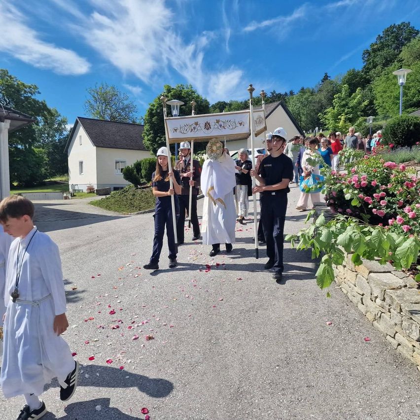 A religious procession is underway on a sunny day. A man in a white robe leads the way, followed by others in uniform and holding banners. The path is lined with flowers, and spectators watch from the side.