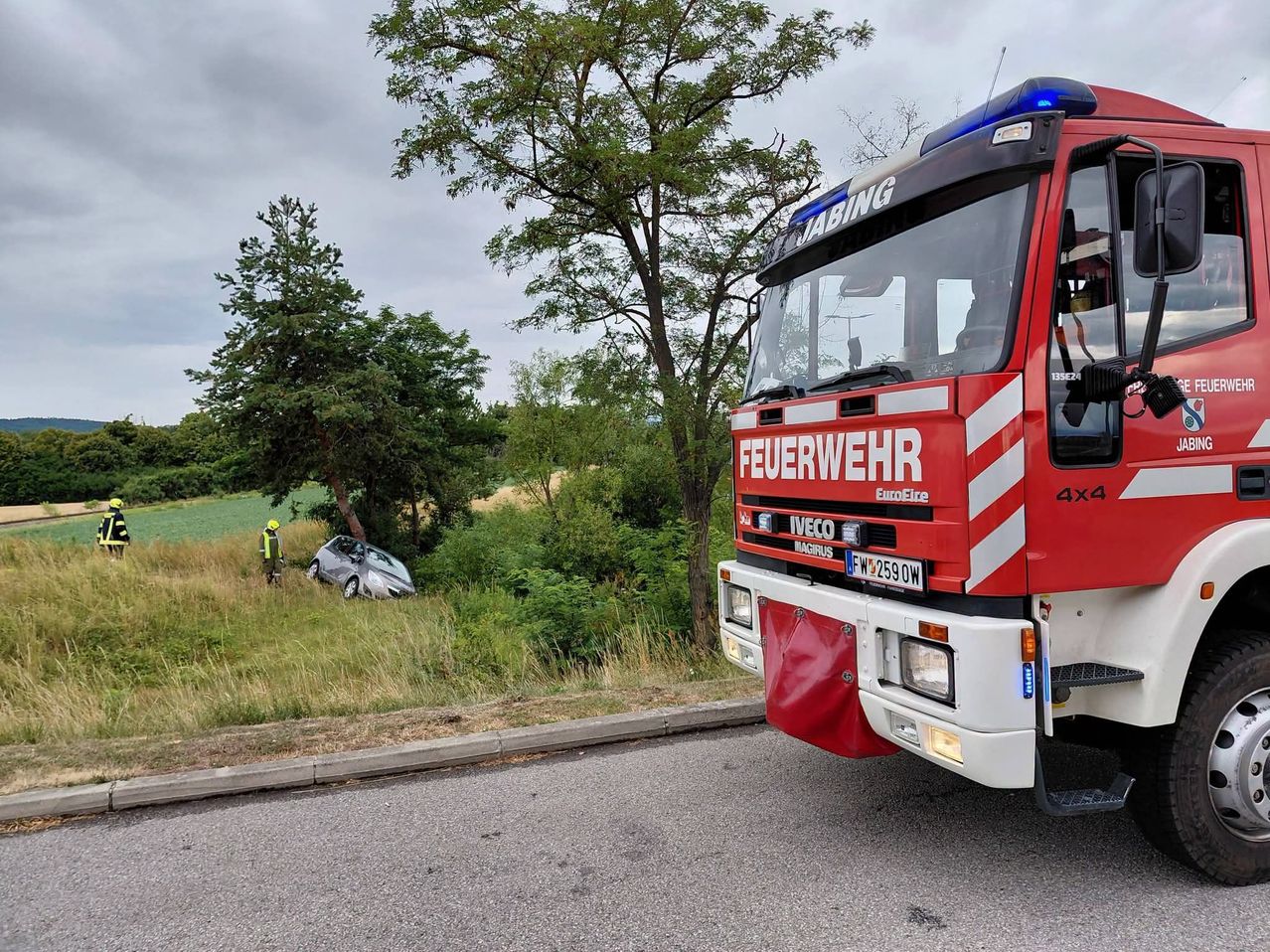 Ein Feuerwehrauto steht am Straßenrand, ein Auto ist ins Gras gecrasht. Eine Person mit gelbem Helm steht daneben.