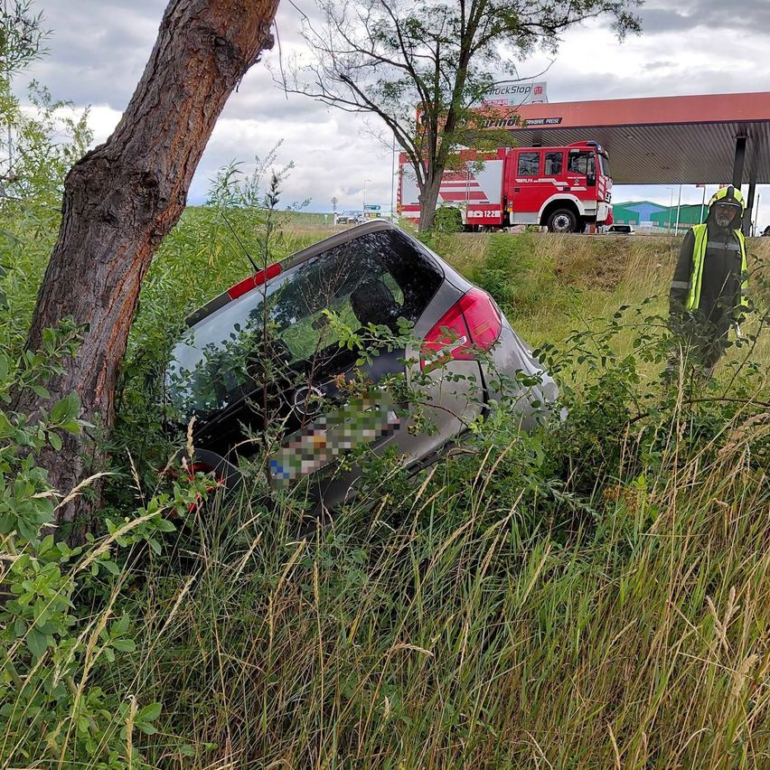Ein demoliertes Auto steckt in Büschen in der Nähe einer Tankstelle. Ein Feuerwehrmann steht in der Nähe mit einem roten Feuerwehrauto im Hintergrund.
