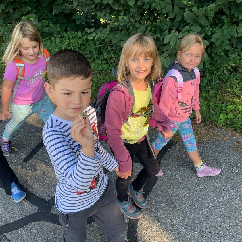 Vier Kinder gehen auf einem Pfad. Ein Junge hält etwas in der Hand. Das Mädchen links trägt einen Rucksack. Hinter ihnen sind Büsche.