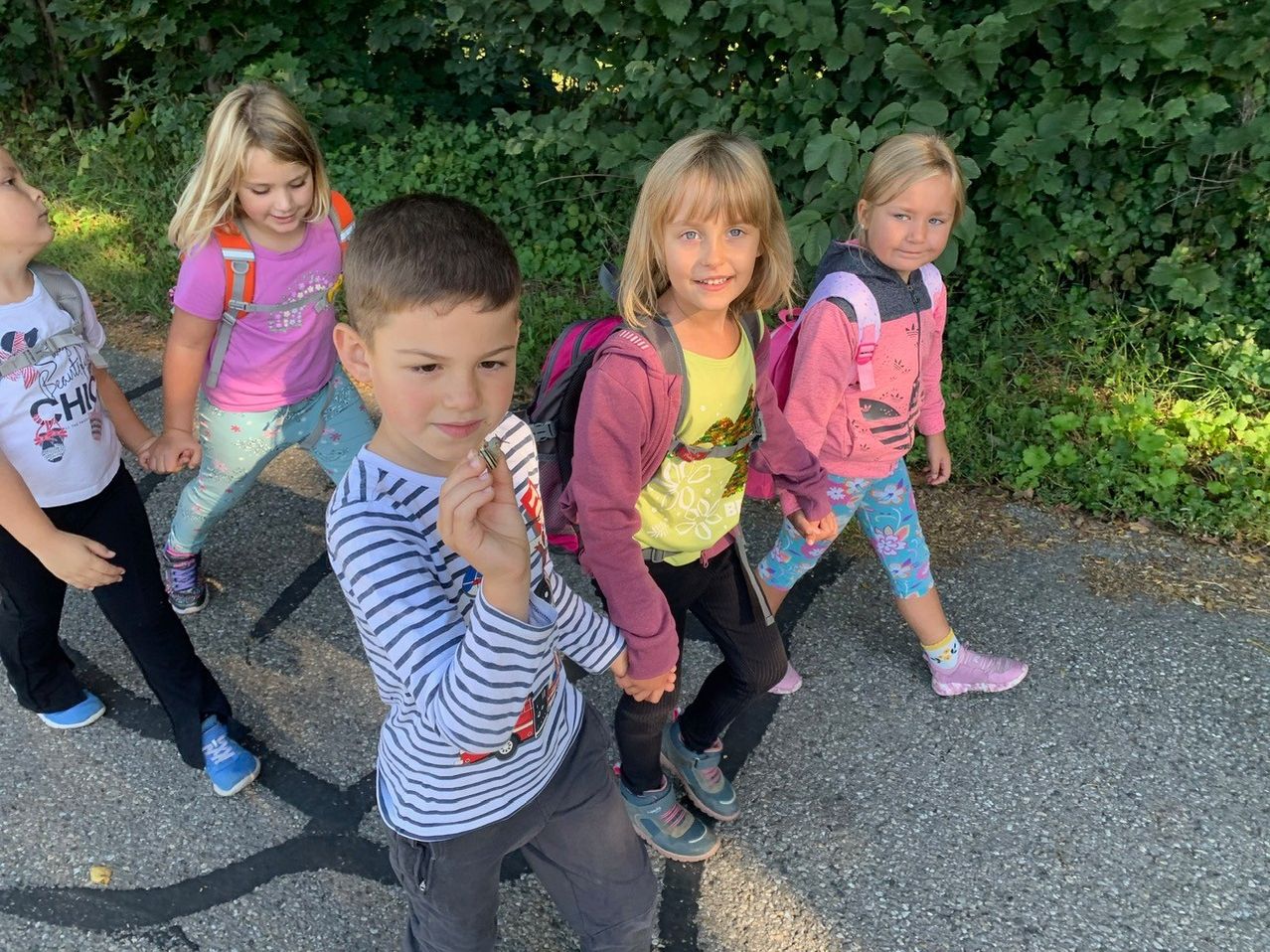 Vier Kinder gehen auf einem Pfad. Ein Junge hält etwas in der Hand. Das Mädchen links trägt einen Rucksack. Hinter ihnen sind Büsche.