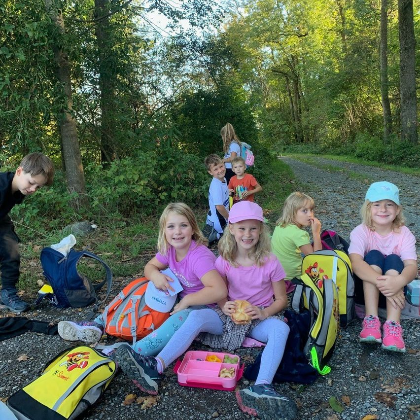 Eine Gruppe von Kindern sitzt auf dem Boden, vielleicht in einem Wald, isst und plaudert. Zwei Mädchen in Pink tragen Hüte. Einige Kinder tragen Turnschuhe. Viele Rucksäcke sind verstreut.