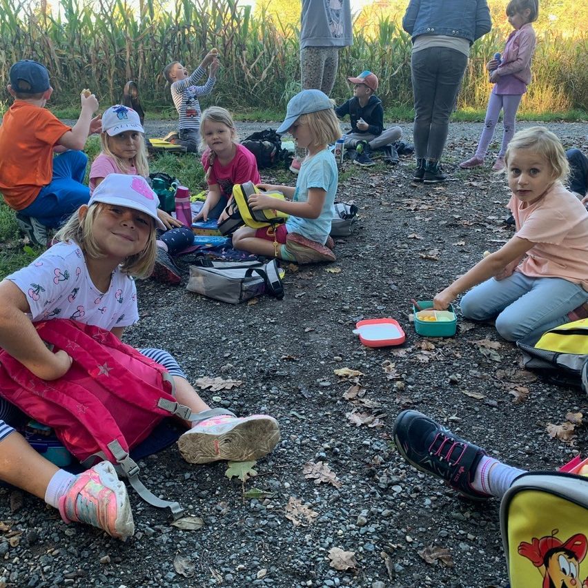 Mehrere Kinder sitzen auf dem Boden, einige mit Mützen, essen und spielen. Einige tragen Rucksäcke und Brotdosen. Einige Erwachsene stehen in der Nähe, einer hält ein Kind. Maispflanzen sind im Hintergrund.