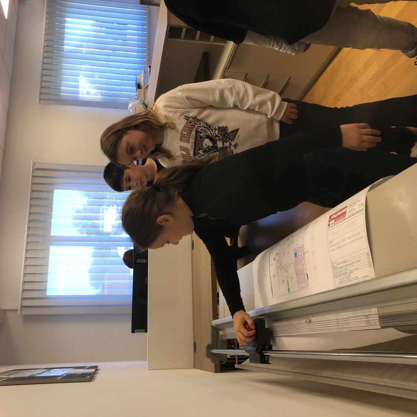 Drei junge Menschen sind in einem Raum, zwei sehen auf ein großes Papier an einer Maschine. Der Raum hat Holzböden, weiße Wände und Fenster mit Jalousien.