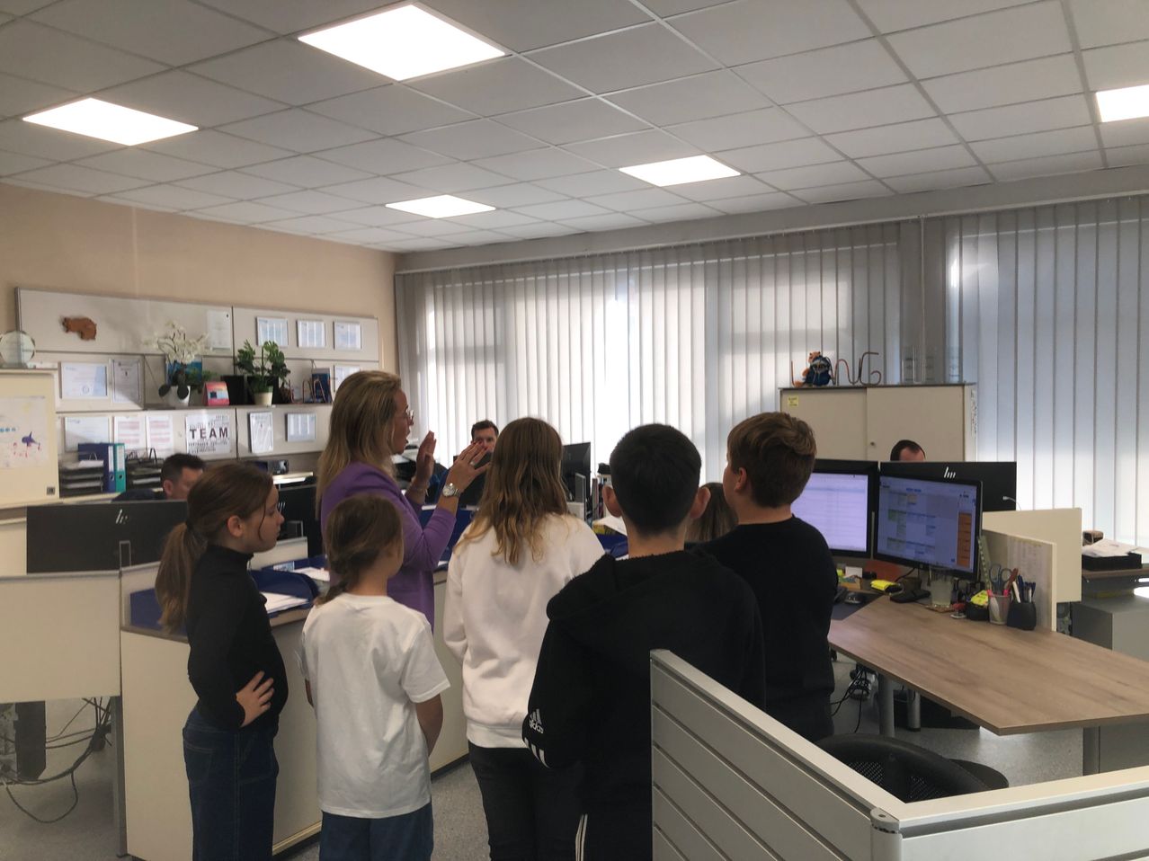 Eine Frau in einem lila Hemd spricht zu einer Gruppe von Kindern in einem Büro mit Computern und Monitoren.